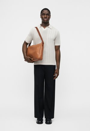 Homme debout portant une chemise blanche à manches courtes en tricot, un pantalon noir à jambes larges, des chaussures noires, tenant un grand sac à bandoulière en cuir marron sur son bras gauche.