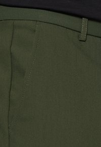 Olivgrüne Hose mit glatter Textur, ausgestattet mit Fronttaschen, geradem Schnitt und dezentem Stitching entlang des Bundes.