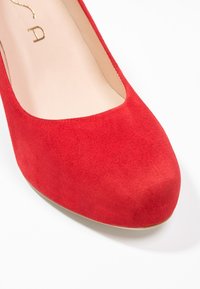 Escarpin en daim rouge avec un bout arrondi, intérieur lisse et un détail subtil de logo doré sur le côté. Semelle plate pour la stabilité.