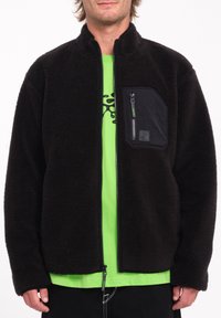 Giacca in pile nera con tasca anteriore con zip, colletto alto e chiusura con zip. Indossata sopra una maglietta verde, realizzata in un tessuto texturizzato.