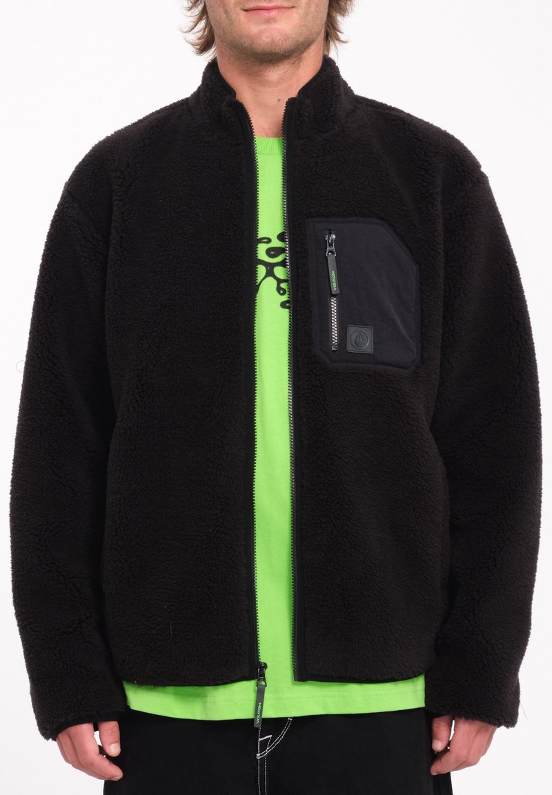 Giacca in pile nera con tasca anteriore con zip, colletto alto e chiusura con zip. Indossata sopra una maglietta verde, realizzata in un tessuto texturizzato.