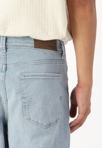 Jeans de mezclilla azul claro con una etiqueta de cuero marrón en la cinturilla, con un diseño de bolsillo trasero y una textura suave.