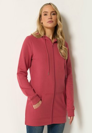 Vrouw met lang blond haar die een donkerroze hoodie met rits en zakken en een blauwe spijkerbroek draagt, staand tegen een effen lichte achtergrond.
