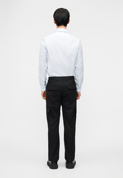 Άνδρας που στέκεται με την πλάτη προς τα εμπρός, φοράει ανοιχτό μπλε πουκάμισο που έχει μπει μέσα σε μαύρο παντελόνι cargo με τσέπες με καπάκια και μαύρα παπούτσια.