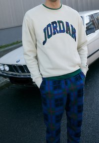 Felpa color crema con la scritta "JORDAN" ricamata a quadri multicolori, abbinata a pantaloni a quadri blu scuro con accenti verdi e rossi.