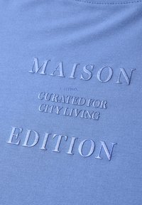 Niebieska tkanina z wytłoczonym tekstem "MAISON EDITION" oraz mniejszym napisem "CURATED FOR CITY LIVING" na środku.
