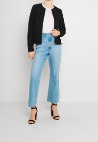 Frau in schwarzer Reißverschlussjacke, weißem Shirt, hellblauen hochtaillierten Jeans und schwarzen High Heels mit Knöchelriemchen steht vor einem einfarbigen Hintergrund.