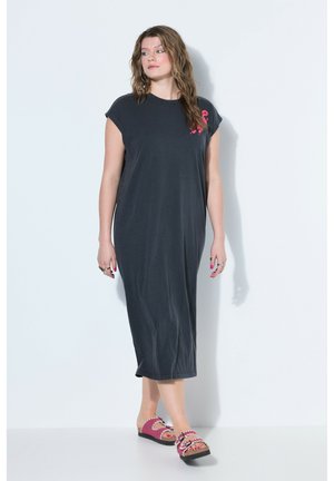 Kvinde med bølget hår iført en sort ærmeløs midi-kjole med et lille pink flamingo-print og pink sandaler, stående mod en hvid baggrund.