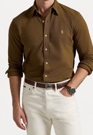 Man draagt een olijfgroen overhemd met lange mouwen opgerold, witte broek, bruine leren riem en een polshorloge met groene wijzerplaat.