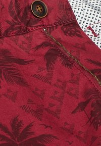 Tissu bourgogne avec un motif de feuilles de palmier et des formes géométriques. Comprend un bouton en laiton et une fermeture éclair visible sur le côté.