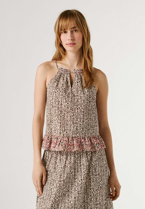 Jonge vrouw met lichtbruin haar, gekleed in een mouwloze beige jurk met geometrische en bloemmotieven, staande tegen een effen neutrale achtergrond.