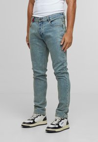 Moški v svetlo modrih slim-fit jeansih, beli majici brez rokavov in črno-belem športnem obutvi stoji ob enobarvni podlagi.