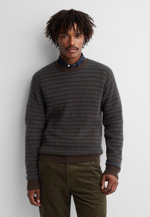 Homme aux cheveux bouclés foncés portant un pull rayé marron et gris par-dessus une chemise sombre et un pantalon en velours côtelé vert olive, debout contre un mur blanc.