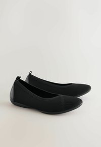 Zapatos planos negros con una parte superior de malla texturizada y acentos en el talón de sintético negro sólido, con punta redondeada y una lengüeta en la parte trasera.