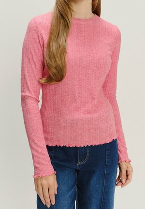 Femme portant un haut en maille rose à manches longues avec des bords festonnés et un jean bleu, debout devant un fond clair uni.