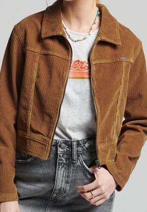 Frau trägt eine kurz geschnittene braune Cordjacke über einem hellgrauen T-Shirt mit Grafik und hoch taillierten, ausgewaschenen schwarzen Jeans, dazu eine Perlenkette.