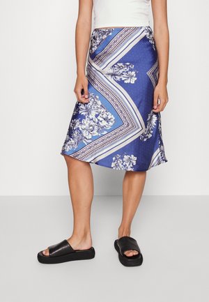 Femme portant une jupe midi bleue à motif floral, un haut blanc sans manches et des sandales noires à enfiler, debout sur un sol gris uni.