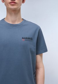 Camiseta de algodón azul con cuello redondo y mangas cortas, con un logotipo en texto del tamaño de un bolsillo en el lado izquierdo del pecho. Textura suave.