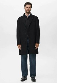 Abrigo largo negro con un diseño entallado, con solapas grandes, delantero de un solo botón, combinado con un suéter negro y jeans azules. Textura suave.