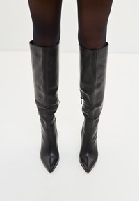 Botas de cuero negro hasta la rodilla con punta afilada, textura suave y cremalleras en los lados internos. Diseño minimalista sin adornos visibles.