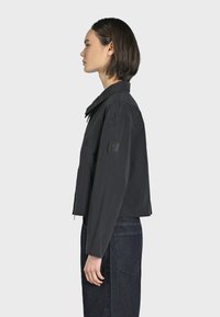 Giacca corta nera con una texture leggera, colletto e una patch sulla manica sinistra. Abbinata a jeans scuri a vita alta.