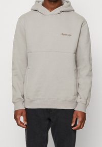 Hellgrauer Kapuzenpullover aus einem weichen, strukturierten Stoff. Mit Kängurutasche und Logo-Stickerei auf der Brust. Rippbündchen an Ärmeln und Saum.