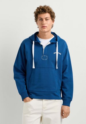 Giovane con capelli ricci che indossa una felpa blu con cordini bianchi e pantaloni bianchi, in piedi con una mano in tasca davanti a uno sfondo semplice.
