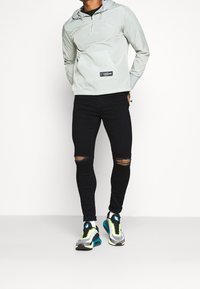Felpa grigio chiaro con cappuccio e tasca frontale, abbinata a jeans skinny neri con strappi al ginocchio e sneakers sportive multicolori.