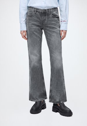 Persona que lleva vaqueros acampanados grises descoloridos, zapatos negros gruesos de plataforma y una camisa de rayas azul claro con texto rojo en el puño.