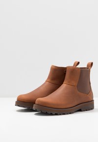 Bottines en cuir brun avec panneaux élastiques sur les côtés et languettes de tirage. Semelles texturées au design robuste pour assurer traction et durabilité.