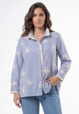 Vrouw draagt een blauw-wit gestreept blouse met knopen, met crèmekleurig bloemenborduursel en kanten rand, gecombineerd met lichtblauwe spijkerbroek.