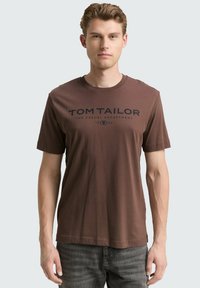 Bruine katoenen t-shirt met korte mouwen en een ronde hals. Voorzien van een donkerblauwe bedrukte logo-tekst over de borst.