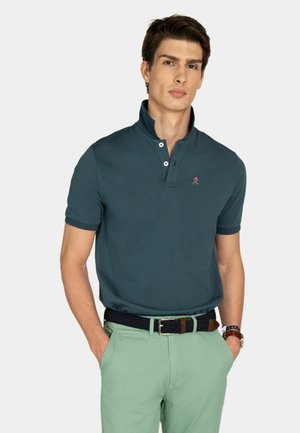 Joven con camiseta tipo polo de manga corta color verde azulado oscuro metida dentro de pantalones verde claro, con un cinturón tejido negro, de pie con las manos en los bolsillos.