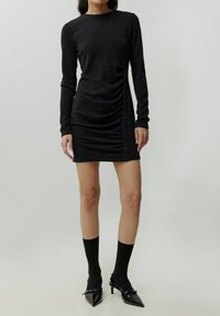 Schwarzes Langarmkleid mit seitlichem Raffung und Knopf-Akzenten, aus weichem Stoff, kombiniert mit schwarzen knielangen Socken und spitzen Absatzschuhen.