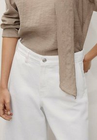 Une personne porte un haut beige à manches longues et un pantalon blanc, tenant sa main sur la taille, dans une pose décontractée et stylée.