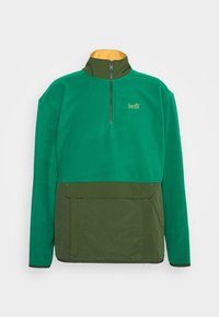 Sudadera de forro polar verde con cuello con cremallera, que presenta una sección inferior en verde oliva y un bolsillo frontal. Logo dorado en el pecho.