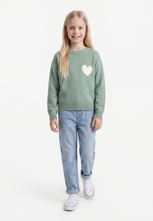 Lächelndes Mädchen mit langen blonden Haaren, das einen grünen Pullover mit weißem Herz, hellblaue Jeans und weiße Sneaker trägt und vor weißem Hintergrund steht.