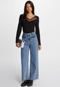 Zwart longsleeve met doorschijnende kanten halslijn, gecombineerd met hooggetailleerde blauwe denim wijdlopende jeans. Model houdt een zilveren clutch vast.