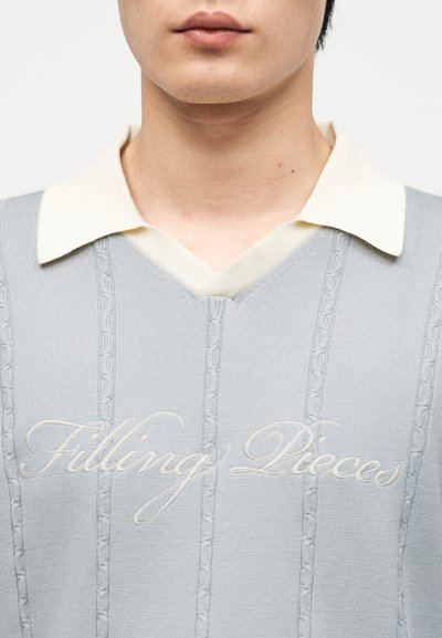 Νεαρό άτομο που φοράει ανοιχτό γκρι πλεκτό πουλόβερ με κάθετα καλωδιακά σχέδια και εκρού γιακά, με το κέντημα "Filling Pieces" στο μπροστινό μέρος.