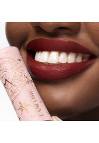 Rouge à lèvres rouge foncé sur des lèvres lisses, avec un tube rose ornée de motifs d'horloge dorés et de détails flocons de neige. Des dents blanches éclatantes sont visibles.