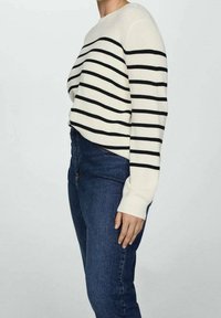 Personne portant un pull crème avec des rayures horizontales noires et un jean bleu foncé taille haute, debout de profil devant un fond uni.