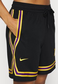 Czarne spodenki sportowe z fioletowymi i żółtymi paskami, elastycznym pasem, sznurkiem do regulacji oraz żółtym logo Nike na dolnej prawej stronie.