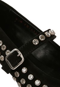 Zapato de ante negro con una correa que presenta herrajes plateados y acentos de strass claros y redondos a lo largo del costado. Forro interior suave.