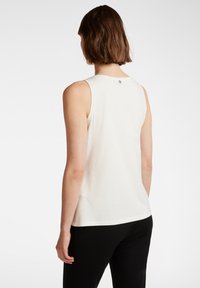 Witte mouwloze top van zacht katoen met een ronde halslijn, met een klein logo op de achterkant; gedragen met zwarte broek.