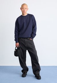 Marineblauwe sweatshirt met geribbelde manchetten, oversized zwarte jeans met rechte pasvorm, en zwarte sneakers; houdt een kleine zwarte tas vast.