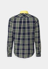 Navy sininen ja vihreä ruudullinen pitkähihaisen paidan, jossa on keltainen kaulus, näkyy takaa valkoista taustaa vasten.