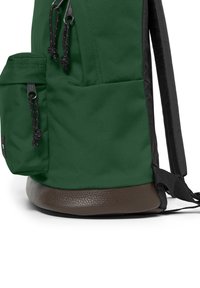 Grön tygryggsäck med en texturerad brun botten, sidoficka med svart dragkedja och justerbara svarta remmar.