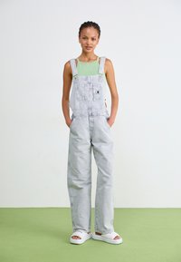 Light gray denim overalls with wide legs, front pocket, and adjustable straps, paired with a green tank top and white slide sandals.