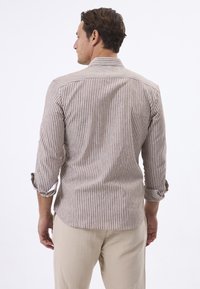 Herenoverhemd met bruine en witte strepen, lichte stof, lange mouwen opgerold, knoopdetails op de manchetten, achterkant gezien. Gecombineerd met beige broeken.