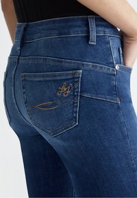 Jeans de mezclilla azul oscuro con una cinturilla alta, que cuentan con bolsillos traseros cosidos con un diseño en espiral en hilo marrón claro.